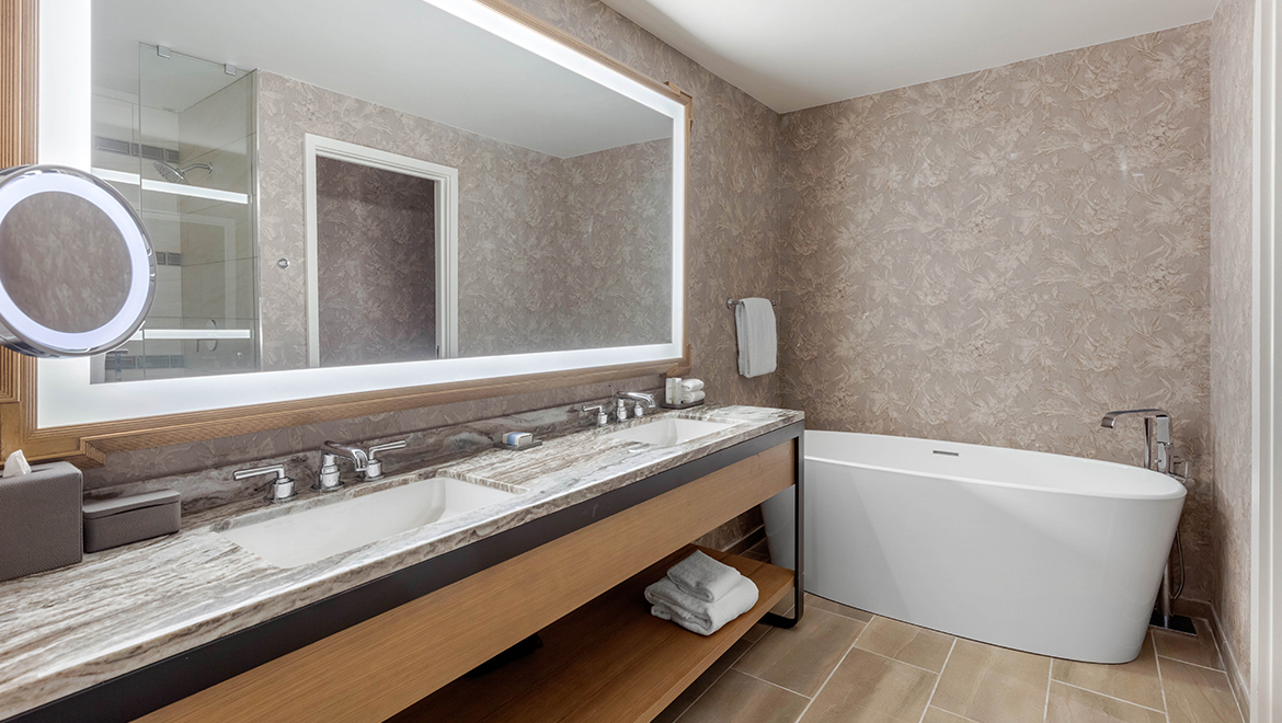 The Champions Executive Suite restroom featuring muted grey wallpaper an oversize mirror, dual sinks and a garden-style bathtub.